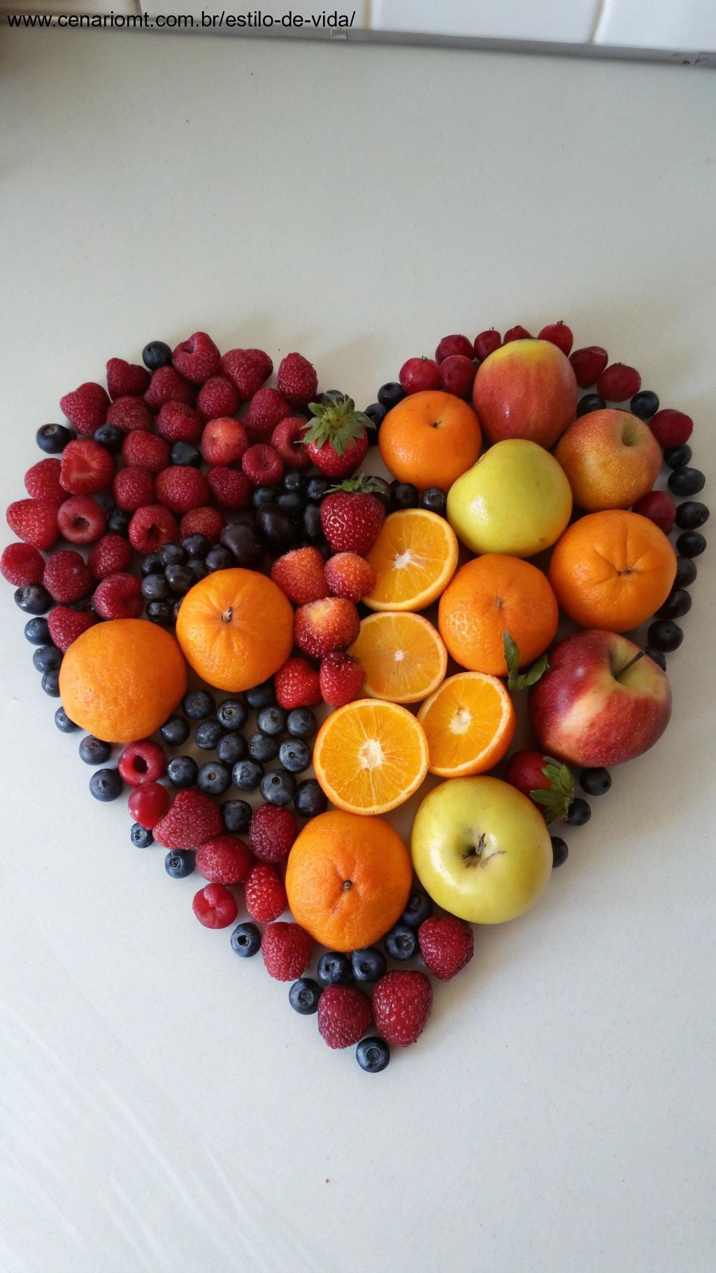 Arranjo de frutas em formato de coração, como frutas vermelhas, laranjas e maçãs.