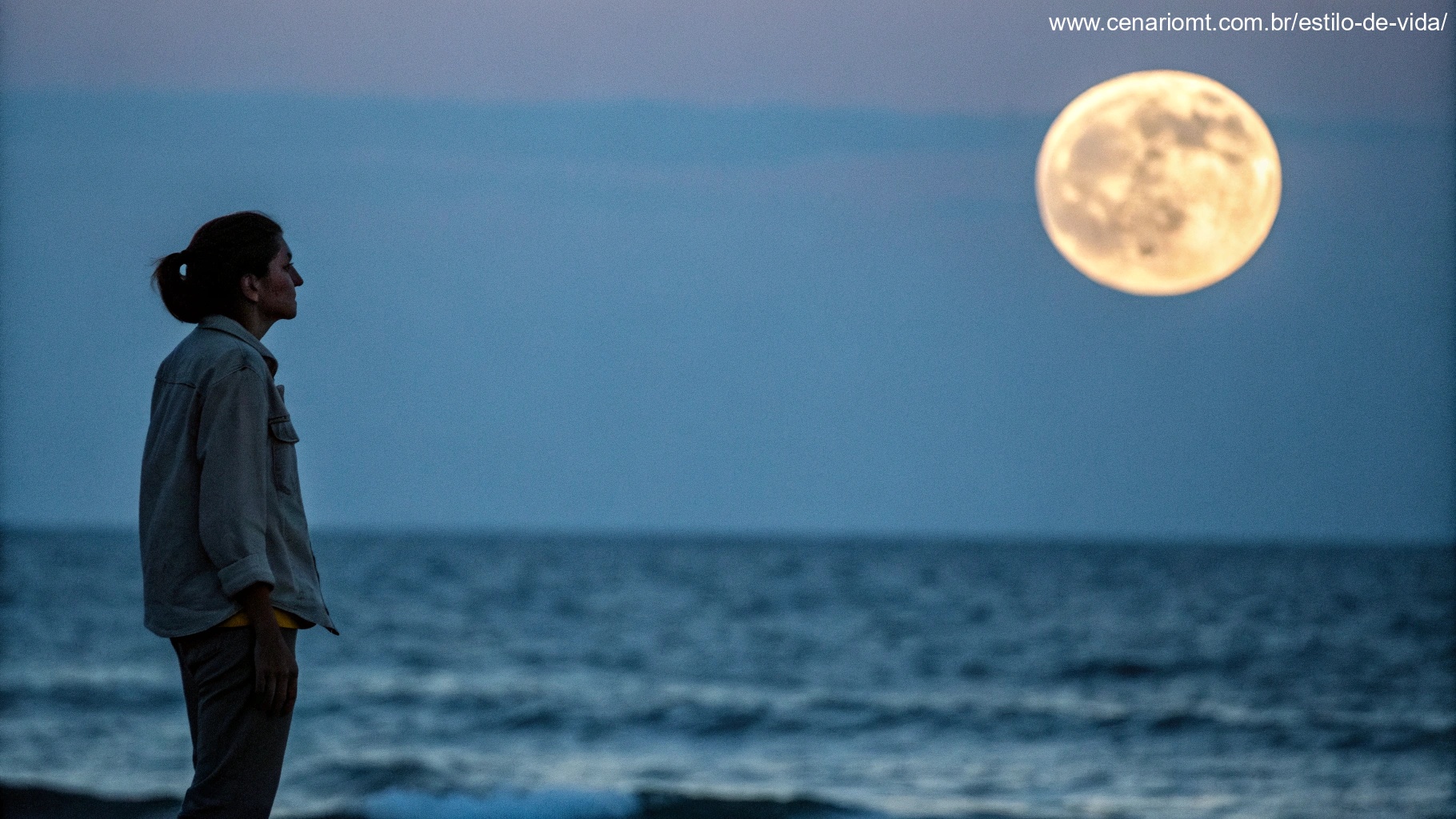Pessoa em pé à beira do oceano sob uma lua minguante, representando a ideia de que a lua minguante influencia as marés e, por isso, afeta o corpo humano