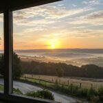 Um amanhecer tranquilo com o sol surgindo sobre colinas e uma janela sugerindo uma manhã acolhedora.