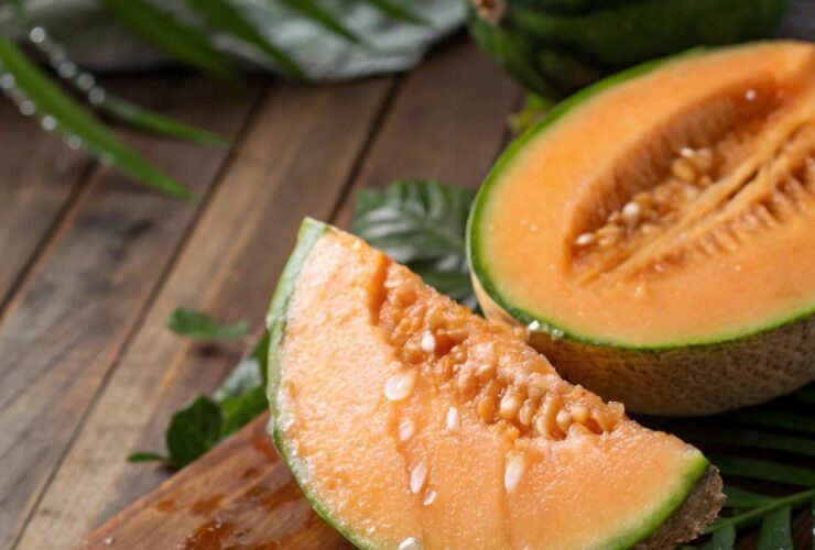 Melão fresco sobre uma mesa de madeira rodeado por folhas tropicais, com gotas de água na superfície, destacando sua carne laranja vibrante.