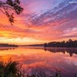 Nascer do sol sereno sobre um lago calmo com cores vibrantes refletindo na água, simbolizando inspiração e energia para uma manhã de sexta-feira.