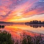 Nascer do sol sereno sobre um lago calmo com cores vibrantes refletindo na água, simbolizando inspiração e energia positiva para a manhã de sábado.