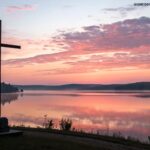 Nascer do sol sereno sobre um lago calmo com uma silhueta de uma cruz em primeiro plano, simbolizando esperança e fé para um novo dia.
