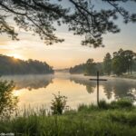 Nascer do sol sereno sobre um lago calmo com uma cruz refletida na água, cercado por vegetação exuberante e luz suave filtrando pelas árvores, evocando paz e inspiração espiritual.