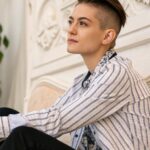 Pessoa com corte de cabelo curto, sentada em uma pose relaxada, com um fundo suave, mostrando elegância e confiança.