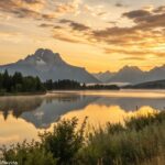 Imagem de um amanhecer sereno sobre uma paisagem calma, com tons dourados suaves no céu refletindo em um lago tranquilo, montanhas ao fundo.