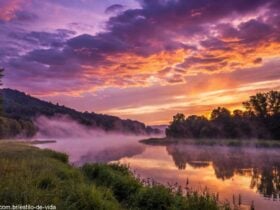 Paisagem serena e mística ao pôr do sol, com tons vibrantes de roxo, laranja e dourado no céu, e um rio calmo refletindo a luz.