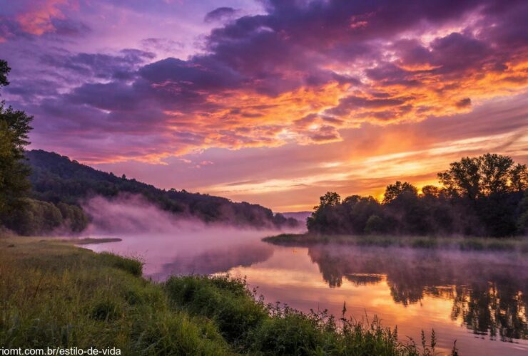 Paisagem serena e mística ao pôr do sol, com tons vibrantes de roxo, laranja e dourado no céu, e um rio calmo refletindo a luz.