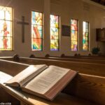 Uma cena serena de manhã de domingo com luz solar passando por vitrais em uma igreja tranquila, um livro de orações aberto em um banco de madeira e um brilho suave de velas no altar.