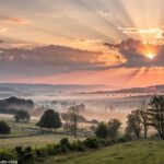 Imagem de um nascer do sol tranquilo sobre uma paisagem rural, simbolizando reflexões espirituais para o dia.