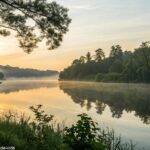 Nascer do sol sereno sobre um lago calmo com uma cruz refletida na água, cercado por vegetação exuberante e luz suave filtrando pelas árvores