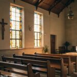 Interior de uma igreja serena com luz suave passando pelos vitrais, bancos de madeira e um altar simples com uma cruz
