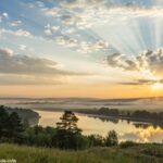 Imagem de uma paisagem tranquila ao amanhecer, com raios suaves de luz e nuvens leves, transmitindo paz e serenidade, ideal para uma mensagem de Bom Dia Abençoado no domingo.