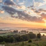 Imagem de um amanhecer sereno sobre uma paisagem tranquila, com nuvens suaves no céu, representando uma manhã esperançosa e abençoada.