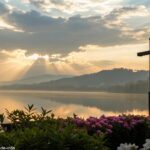 Nascer do sol sereno sobre um lago calmo com uma silhueta de cruz em primeiro plano, cercado por flores desabrochando e raios de luz suaves atravessando as nuvens
