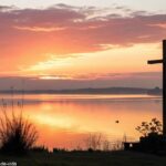 Nascer do sol sereno sobre um lago calmo com uma silhueta de cruz em primeiro plano, envolto em luz dourada suave e uma atmosfera pacífica