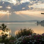 Um nascer do sol sereno sobre um lago calmo com uma silhueta de cruz em primeiro plano, cercado por flores desabrochando e raios de luz suaves atravessando as nuvens