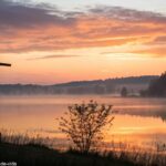 Nascer do sol sereno sobre um lago calmo com uma silhueta de uma cruz em primeiro plano, cercado por luz suave e neblina