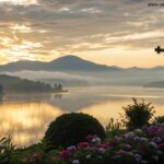Nascer do sol sereno sobre um lago tranquilo com uma silhueta de cruz em primeiro plano, luz dourada refletindo na água, cercado por montanhas nebulosas e florescendo