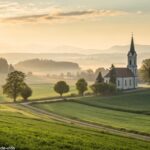 Paisagem tranquila de manhã com luz suave iluminando uma igreja distante, cercada por campos verdes, simbolizando uma manhã calma e abençoada de terça-feira para um grupo religioso.