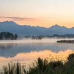 Uma imagem de um amanhecer tranquilo sobre um lago calmo, com névoa suave na água e montanhas ao fundo, representando o início de um novo dia e a oportunidade para sabedoria.