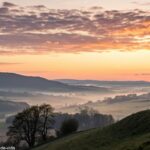 Imagem serena de um amanhecer no campo com nuvens suaves e uma silhueta de cruz no topo de uma colina, evocando paz e espiritualidade para um momento de oração católica e evangélica.
