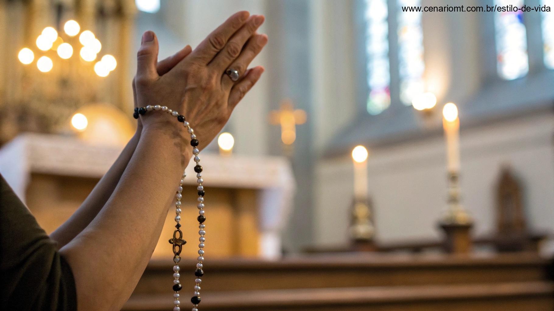 Mãos em posição de oração, com um terço sobre elas, em um fundo desfocado de um altar de igreja e luz suave de velas