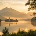 Paisagem serena ao amanhecer com luz dourada iluminando um lago calmo, rodeado por árvores e montanhas ao fundo, simbolizando paz e reflexão espiritual.