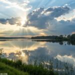 Paisagem tranquila com um lago calmo refletindo o céu, com luz suave do sol entre as nuvens, representando um momento de reflexão e oração para a manhã de quinta-feira.