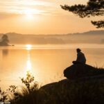 Cena tranquila de um nascer do sol sobre um lago sereno, com uma pessoa lendo a Bíblia.