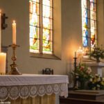 Imagem serena e tranquila de um altar de igreja com velas acesas e uma luz suave entrando pelas janelas de vitral, simbolizando reflexão e oração para o Santo do Dia.