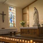 Imagem de uma capela católica tranquila, com um altar de madeira e uma estátua de santo. A cena tem um ambiente reflexivo com luz suave entrando pelas janelas de vitrais.