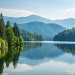 Uma cena tranquila da natureza com um lago calmo refletindo o céu claro, rodeado por árvores verdes e montanhas ao fundo.