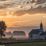 Amanhecer tranquilo sobre uma igreja, com luz suave e nuvens serenas, representando a manhã de sexta-feira para grupos religiosos refletirem e orarem.