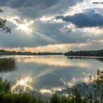 Paisagem tranquila com luz suave atravessando as nuvens sobre um lago sereno.