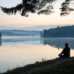 Imagem de um cenário sereno com um lago calmo e uma pessoa meditando ao amanhecer.