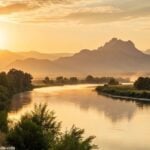 Paisagem tranquila ao amanhecer, com luz dourada suave iluminando um rio calmo e montanhas ao fundo, simbolizando reflexão e paz espiritual.