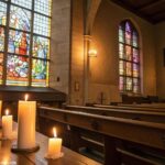 Cena de igreja tranquila com vitrais e velas, refletindo luz suave nos bancos.