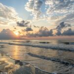 Imagem de um amanhecer tranquilo sobre um cenário sereno, com nuvens suaves no céu e ondas suaves batendo na praia, evocando uma sensação de calma e espiritualidade.