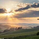 Imagem de um nascer do sol sobre o campo com uma cruz luminosa no céu, representando fé e esperança