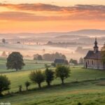 Paisagem tranquila com nascer do sol e uma igreja simples ao fundo, representando uma manhã de fé