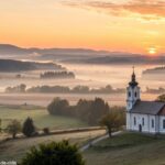 Imagem de uma manhã tranquila com o nascer do sol e símbolos religiosos ao fundo.
