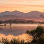 Uma imagem de um amanhecer tranquilo sobre um lago calmo, com luz suave refletindo na água e montanhas ao fundo, criando uma sensação positiva.
