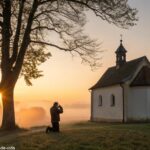 Imagem de uma capela ao amanhecer com uma pessoa orando sob uma árvore