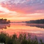 Imagem de um amanhecer sereno sobre um lago calmo, com o céu em tons suaves de rosa e laranja.