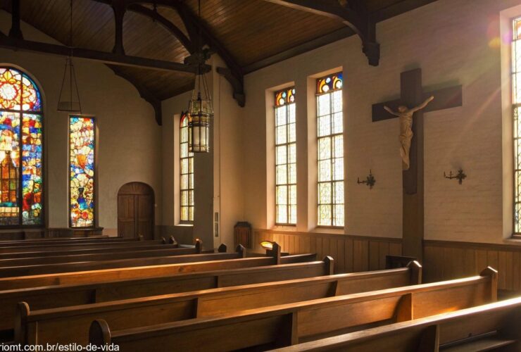 Igreja vazia com luz do sol entrando pelos vitrais, iluminando uma cruz de madeira simples no altar, atmosfera tranquila e reflexiva