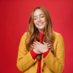 girl feeling like heart warming as remembering sweet tender memories with closed eyes silly broad smile holding hands chest pressing soul posing nostalgic touched red background min 1