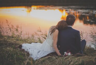 Casal de noivos sentado na grama verde na frente do corpo de água ao pôr do sol - Fotos do Canva
