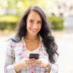 A smiling brunette is writing messages on her mobile phone. A young woman reads received messages and writes back on a smartphone. - Fotos do Canva
