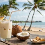 Imagem de uma batida de coco tropical em um copo, decorada com flocos de coco e um canudo, com fundo de palmeiras e praia.